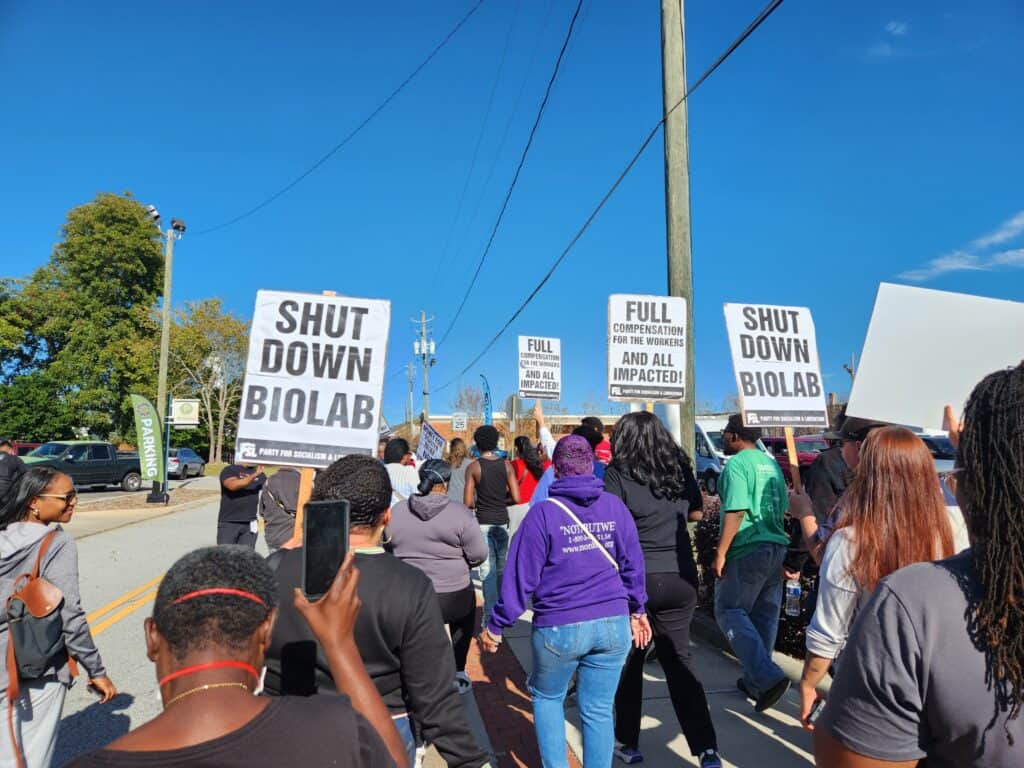 A group of people march down a street while holding signs which read "SHUT DOWN BIOLAB" and other messages.